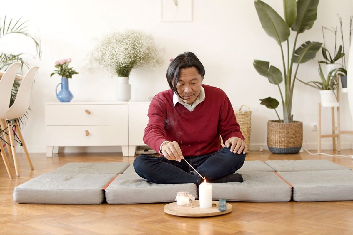À propos Asian man sitting on mat lighting incense with candle in serene living room. Cozy and calm atmosphere.